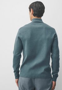 Pull turquoise avec un col montant, des manches longues et une texture lisse. Le dos présente un design ajusté et des poignets côtelés.