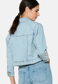 Helehelesin sinine teksajakk, mis on lühikese lõikega, pika varrukaga ja nupuga mansettidega. Omab tagakotisid ja minimaalset õmblusdetaili.