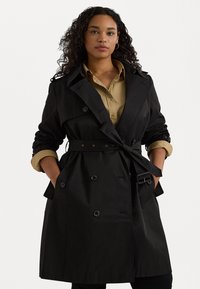 Svart trenchcoat med bälte i midjan, dubbelknäppt knappslå och uppkavlade ärmar över en khaki skjorta. Slitstarkt och mjukt tyg.