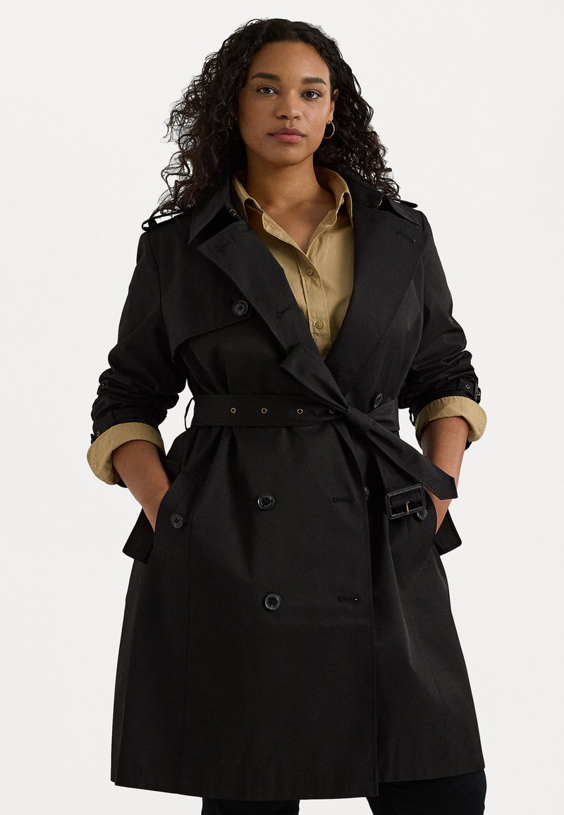 Svart trenchcoat med bälte i midjan, dubbelknäppt knappslå och uppkavlade ärmar över en khaki skjorta. Slitstarkt och mjukt tyg.