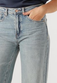 Lichtblauwe denim jeans met een hoge taille, rechte pijpen en zichtbare slijtage langs de dijen. Enkele voorzak zichtbaar.