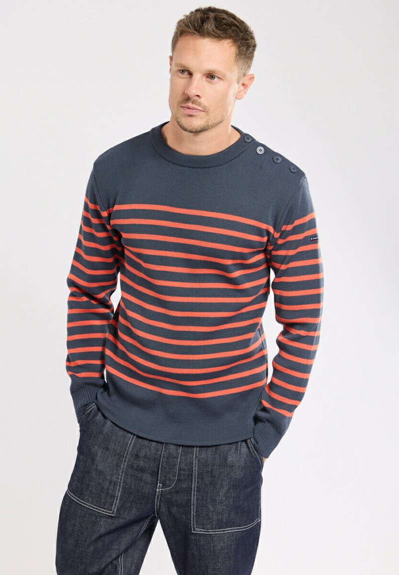 Pull marine bleu avec des rayures horizontales rouges, col rond, manches ajustées, et quatre boutons décoratifs sur l'épaule gauche.