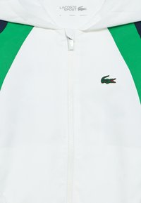 Biela bunda so zelenými a námorníckymi bočnými panelmi, predný zips, textúrovaná tkanina, s malým zeleným logom krokodíla na hrudi.