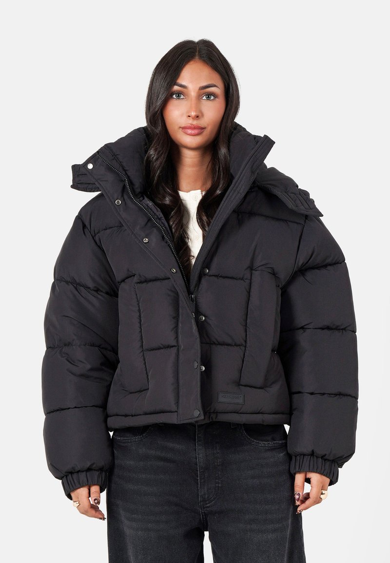 Kvinna iklädd en svart kort puffjacka med hög krage och svartajeans, som står mot en enfärgad ljus bakgrund.
