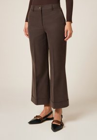 Pantalones de pierna ancha marrón oscuro con una textura suave, que presentan un cierre frontal con botón y dobladillos enrollados. Combinados con unas bailarinas negras de punta afilada.