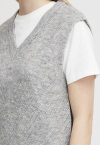 Grau gestrickte Weste mit einem strukturierten Rautenmuster, ausgestattet mit einem V-Ausschnitt und ärmellosem Design über einem weißen T-Shirt. Weiches, wollähnliches Material.