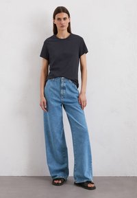 T-shirt grigio scuro con maniche corte, abbinato a jeans in denim a gamba larga di un blu chiaro e infradito neri. Tessuto dalla consistenza liscia, design casual.