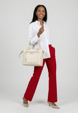 Mujer sonriente de pie, vestida con blusa blanca, pantalones rojos y tacones beige, sosteniendo un bolso grande beige con bolsillo con cremallera frente a un fondo liso.