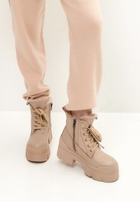 Botas de tobillo beige con cordones y suela gruesa, que cuentan con cremalleras y un borde de piel de pelo suave para mayor comodidad y detalle. Textura de cuero suave.