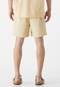 Svetložlté šortky s voľným strihom, vyrobené z textúrovaného materiálu, zobrazené zozadu. V kombinácii s hnedými šľapkami.