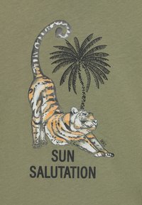 Olivgrüner Stoff mit einem sich streckenden Tiger, akzentuiert mit orange- und schwarzen Streifen. Oben ist eine Palme und der Text "SONNENGRUSS".