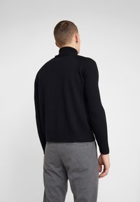 Man in een zwart ribbeltrui met lange mouwen en een grijze broek, staand met de rug naar de camera tegen een effen lichte achtergrond.
