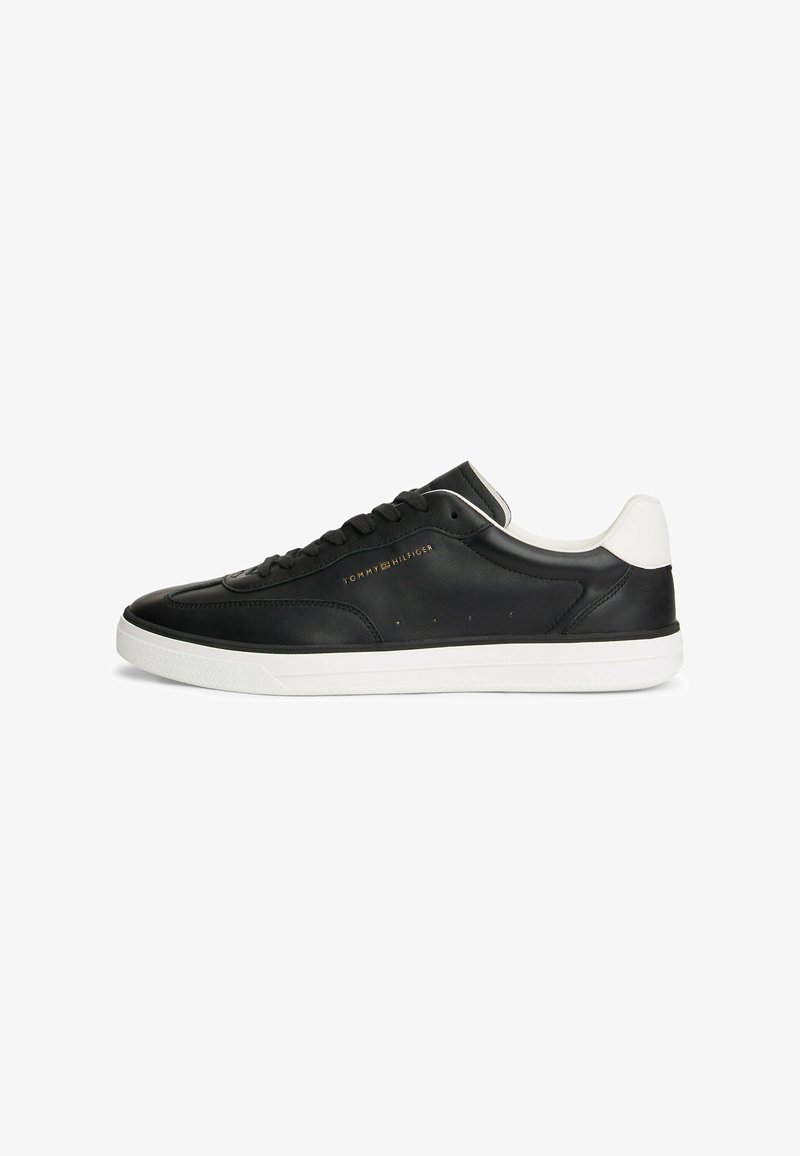 Baskets basses noires avec semelle blanche et languette de talon, lacets noirs, et logo doré « Tommy Hilfiger » sur le côté.