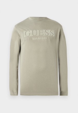 Maglietta a maniche lunghe in cotone beige chiaro con logo testurizzato "GUESS EST. 1981" sul davanti e design a girocollo. Texture liscia, vestibilità casual.
