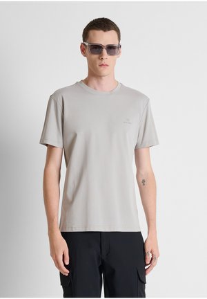 Giovane uomo con una maglietta grigio chiaro, pantaloni cargo neri e occhiali da sole scuri, in piedi davanti a uno sfondo bianco uniforme.