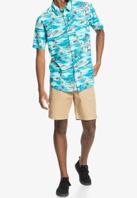 Turkoosi, lyhythihainen paita, jossa rantakohtauskuvio, mukaan lukien palmupuut ja purjelaivat, yhdistettynä beigenvärisiin shortseihin ja mustiin kenkiin.