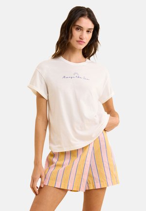 T-shirt en coton blanc avec des manches retroussées, portant "Always the Sun" brodé en bleu au-dessus d'un graphique de soleil. Porté avec un short rayé jaune-rose.