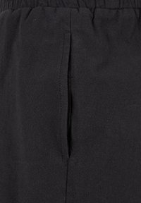 Gros plan sur un tissu noir montrant une poche à fente verticale avec des coutures visibles et une ceinture élastique au-dessus.