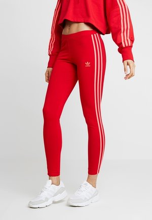 Červené sportovní legíny vyrobené z pružného materiálu, s třemi vertikálními světle červenými pruhy po stranách a malým logem nad kotníkem.