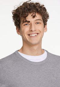 Grå tröja med rund halsringning och subtil textur, bärs över en vit T-shirt. Inkluderar en liten broderad logotyp på bröstet.