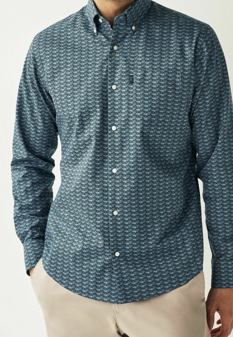Homme portant une chemise bleue à boutons avec un petit motif sombre de voitures et un pantalon beige, debout les mains dans les poches.