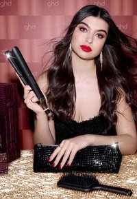 Zwarte keramische haarstijltool vastgehouden in een hand, gouden glinsterende achtergrond met een gestructureerde zwarte clutch en een zwarte haarborstel in de buurt.