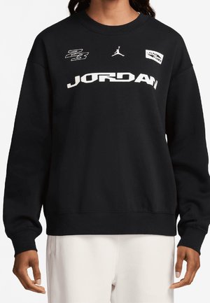Schwarzer Sweatshirt mit weißem "JORDAN"-Schriftzug, Jumpman-Logo und zwei kleinen Grafik-Patches, getragen von einer Person, deren Hände unterhalb der Taille sichtbar sind.