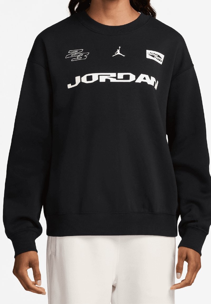 Sweat-shirt noir avec texte blanc "JORDAN", logo Jumpman et deux petites pièces graphiques porté par une personne avec les mains visibles sous la taille.