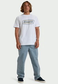 Νεαρός άνδρας με καστανά μαλλιά μήκους ώμου, φορώντας ένα λευκό T-shirt Billabong, ανοιχτό μπλε τζιν και μαύρα αθλητικά παπούτσια, στέκεται όρθιος.