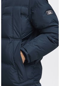 Navy puffer jacket featuring quilted sections, a zippered pocket, and ribbed cuffs. Fabric is shiny with a smooth texture.