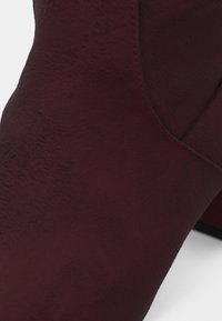 Gros plan d’une botte cheville en daim bordeaux foncé avec une couture courbe lisse et un talon bloc sur fond blanc.