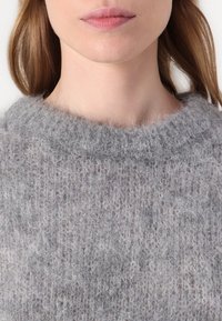 Világosszürke pulóver, kötött, puha, bolyhos textúrával és széles, kör alakú nyakkivágással. Az anyag szemmel láthatóan laza szövésmintával rendelkezik.