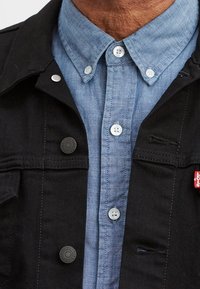 Musta farkkutakki, jossa on hopeiset nepparit, vaaleansinisen chambray-paidan päällä, jossa on kaulus ja nappisulku. Punainen label takin taskussa.