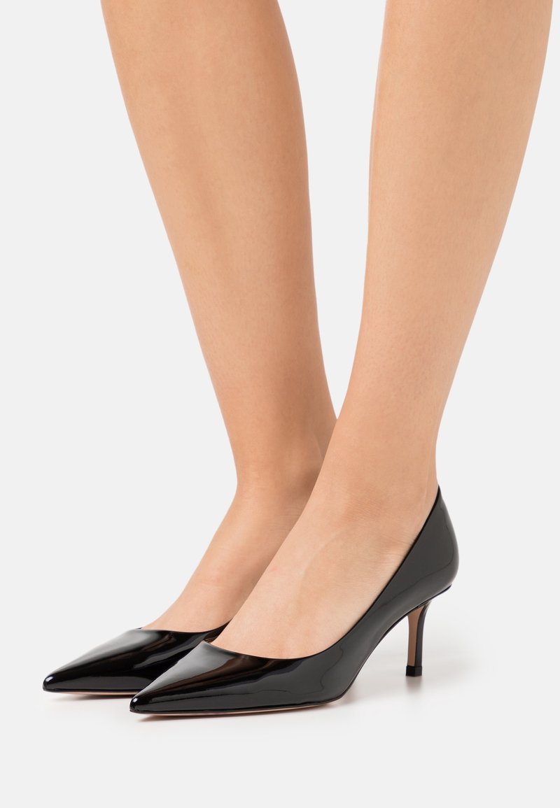 Jambes portant des escarpins noirs brillants à bout pointu avec un petit talon fin sur un fond blanc uni.