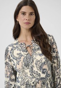 Bluse med blomsterprint i crème, med blåt paisleymønster. Nøglehulslinning med bånddetalje og blafrende ærmer. Blødt stof.