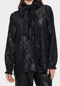 Svart genomskinlig blus med ett texturerat mönster, knytkrage och draperade manschetter, tillsammans med svarta läderbyxor.