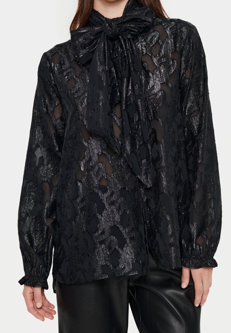 Svart genomskinlig blus med ett texturerat mönster, knytkrage och draperade manschetter, tillsammans med svarta läderbyxor.