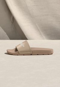 Μπεζ πλατφόρμα σανδάλι με φαρδιά λωρίδα που φέρει λευκό λογότυπο κειμένου, τοποθετημένη σε ουδέτερη επιφάνεια με απαλό υφασμάτινο φόντο.