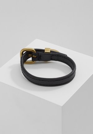 Czarna skórzana opaska na nadgarstek z klamrą i pętlą w kolorze złotym, wyłożona w zwiniętej formie na białej geometrycznej podstawie na tle w kolorze szarym.