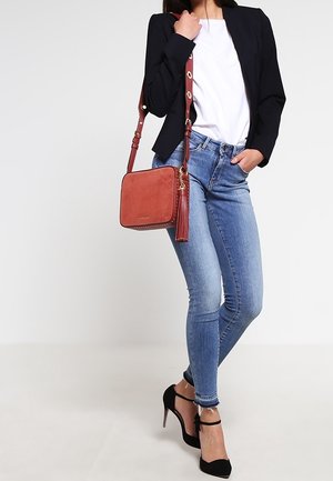 Rød ruskinds crossbody taske med en struktureret overflade og en frynseaccent, kombineret med blå skinny jeans og en tætsiddende sort blazer over en hvid top.