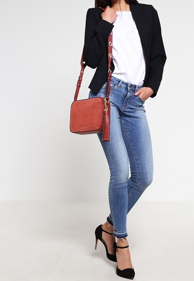 Rode suède crossbody tas met een gestructureerd oppervlak en een kwastaccent, gecombineerd met blauwe skinny jeans en een getailleerde zwarte blazer over een witte top.