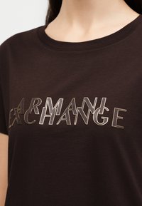 Gros plan d'une personne portant un t-shirt brun foncé avec des lettres blanches et dorées en chevauchement épelant "ARMANI EXCHANGE" sur la poitrine.