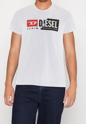 Mężczyzna w białej koszulce marki Diesel z czerwonym i czarnym graficznym napisem, zestawionej z ciemnoniebieskimi dżinsami, stoi na białym tle.