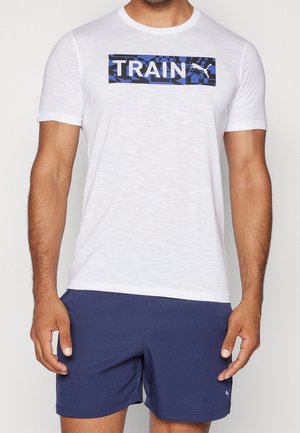 Mężczyzna ubrany w białą koszulę z krótkim rękawem z niebieskim wzorem i napisem "TRAIN" oraz granatowe spodenki, stojący z luźno opuszczonymi ramionami.