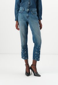 Jeans din denim, cu glezna conică, de culoare albastră decolorată, ornamentate cu broderie florală albă la tiv, asortate cu pantofi cu toc înalt negri.