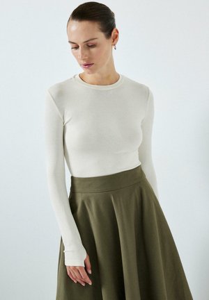 Kvinde med mørkt hår iført en hvid langærmet top og olivengrøn højhåndet nederdel, kigger nedad med en neutral baggrund.