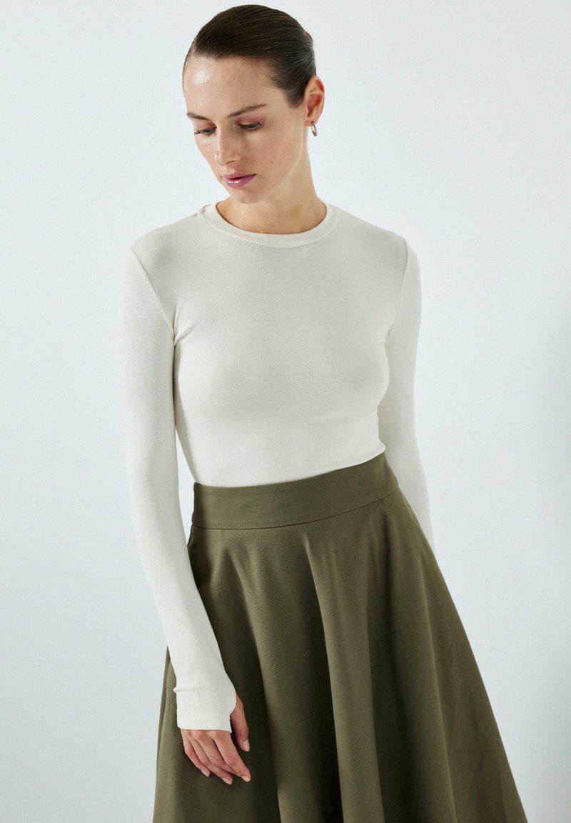 Femme aux cheveux foncés portant un haut blanc à manches longues et une jupe taille haute vert olive, regardant vers le bas avec un fond neutre.