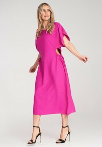 Fuchsia Midi-Kleid mit kurzen Ärmeln, seitlichen Ausschnitten und Kordelzugakzenten an der Taille, kombiniert mit schwarzen, trägerlosen High-Heels.