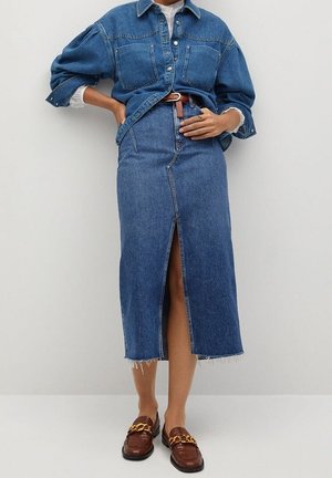 Vrouw draagt een dichtgeknopte spijkerjas, een spijkerrok met hoge taille en een split aan de voorkant, een bruine riem en bruine loafers met gouden kettingen.