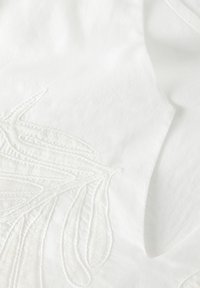 Tissu en coton blanc avec une broderie blanche complexe, présentant des motifs floraux et des points en relief, mettant en valeur la profondeur et le détail.